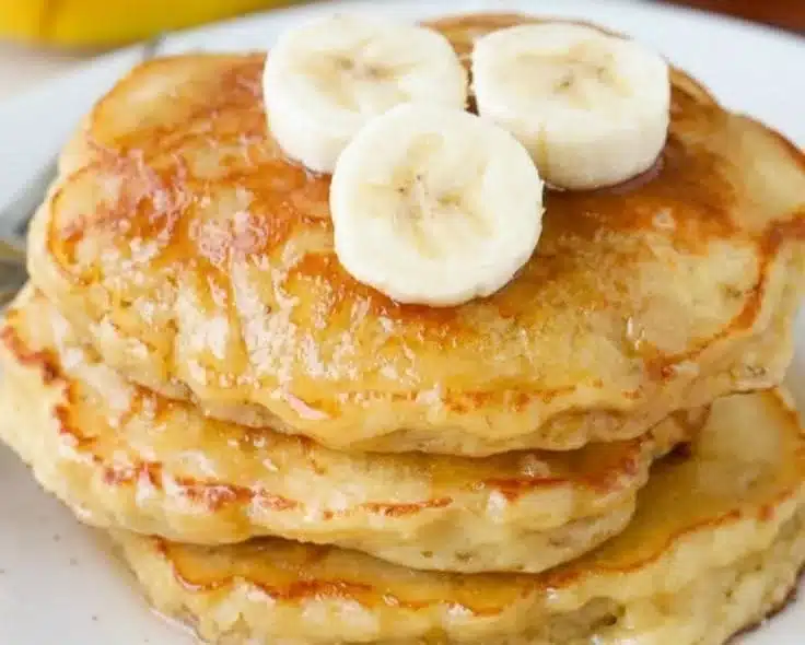 Delicious banana pancakes stacked on a plate with syrup and bananas